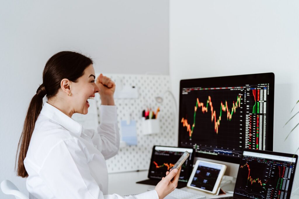 Happy excited young female cryptocurrency trader sitting at desk looking at multiple screens