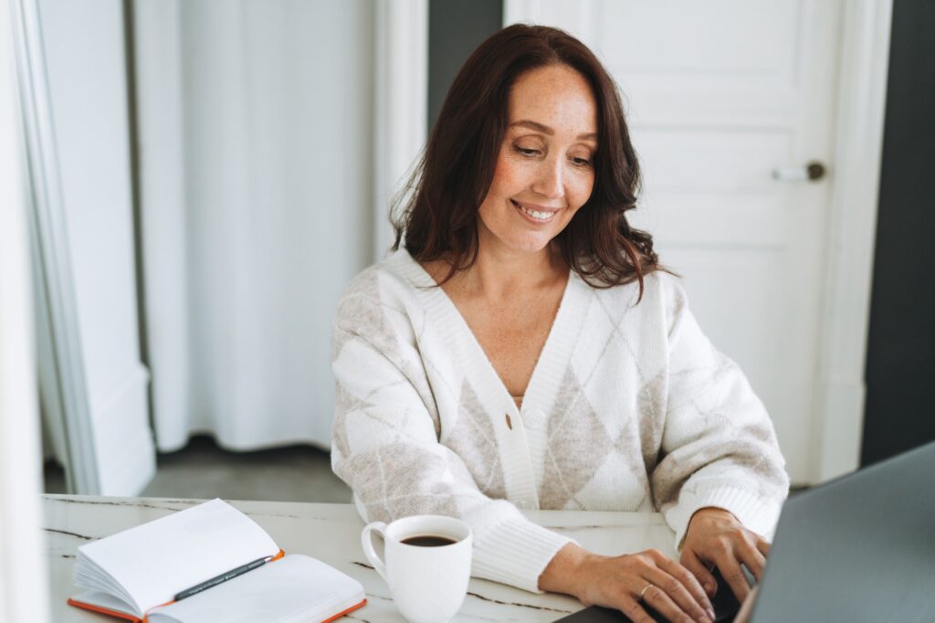 Smiling brunette woman with long hair in white cardigan working on laptop in bright modern office