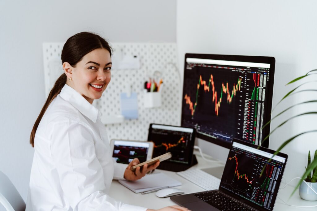 Young girl cryptocurrency trader sitting at desk with multiple screens looking at camera